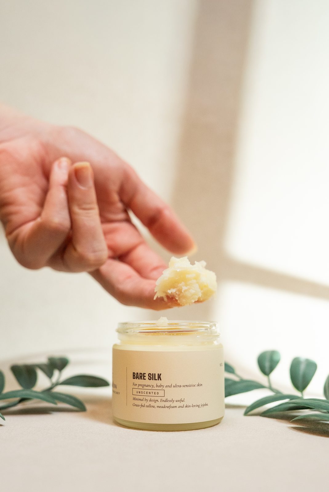 A fingertip lifting a scoop of Bare Silk Unscented from the open jar